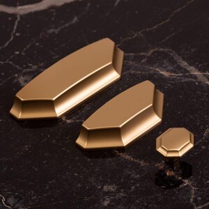 Puede incluir: Juego de herrajes dorados para armarios, que incluye dos tiradores alargados y un pomo. Los herrajes tienen un diseño facetado y un acabado mate. Los artículos están dispuestos sobre una superficie oscura veteada.