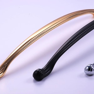 Könnte beinhalten: Drei gebogene Schrankgriffe in Gold, Schwarz und Silber. Der goldene Griff befindet sich links, der schwarze in der Mitte und der silberne rechts. Jeder Griff hat ein abgerundetes Ende und ein gerilltes Design.