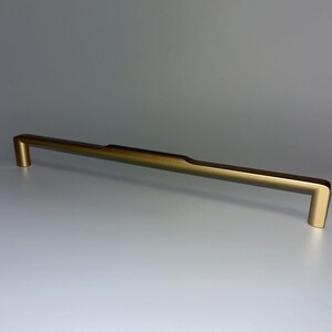 Può includere: Una maniglia per mobili color oro con una barra rettangolare e estremità ad angolo retto. La maniglia ha una sottile scanalatura vicino al centro. L'hardware è progettato per una facile installazione e aggiunge un tocco moderno.
