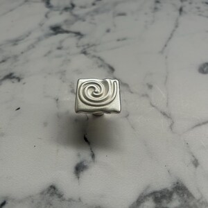 Op de afbeelding: Een vierkante, zilverkleurige kastknop met een verhoogd spiraalontwerp. De knop is gecentreerd op een wit en grijs marmerpatroon oppervlak. De knop is waarschijnlijk van metaal.