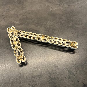 Könnte beinhalten: Zwei längliche Schubladengriffe aus Metall mit cremefarbener Oberfläche und goldfarbenen Akzenten. Jeder Griff zeigt ein symmetrisches, durchbrochenes Schnörkelmuster entlang seiner Länge, das durch sich wiederholende blattartige Formen geprägt ist.