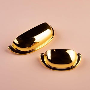 Può includere: Due maniglie dorate per mobili a forma di mezza luna. Le maniglie hanno un design curvo con piccole maniglie arrotondate a ciascuna estremità. Sono poste su uno sfondo rosa tenue, creando un contrasto con l'oro brillante.