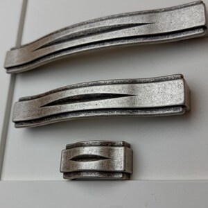 Pode incluir: Três puxadores de armário de metal em tom prateado de diferentes comprimentos dispostos verticalmente. Cada puxador apresenta um design retangular curvo com uma ranhura horizontal central que se estreita nas extremidades, com acabamento escovado.