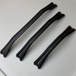 Peut inclure: Trois poignées de meubles noires au design incurvé et décoratif. Les poignées sont disposées en diagonale sur une surface blanche. Chaque poignée a un centre nervuré et des extrémités évasées, suggérant un style vintage ou traditionnel.
