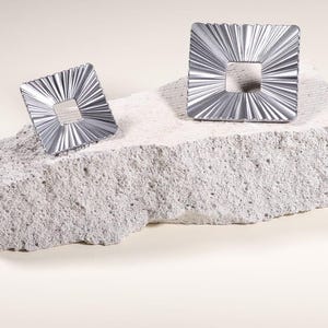 Può includere: Due oggetti decorativi quadrati color argento con un'apertura quadrata centrale e un design a raggi. Sono esposti su una superficie di pietra ruvida grigio chiaro su uno sfondo neutro.