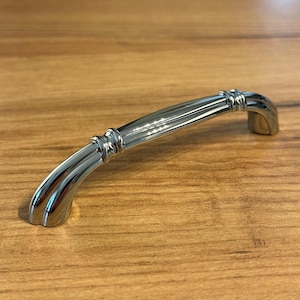Puede incluir: Un tirador de armario de cromo pulido con un diseño curvo y arqueado. El mango presenta un acabado metálico liso y reflectante, con dos acentos decorativos de anillos estriados situados cerca de cada extremo de la barra.