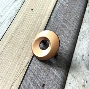 Op de afbeelding: Een bronskleurige kastknop met een cirkelvormig ontwerp en een centraal gat. De knop heeft een gladde, metallic afwerking en is geplaatst tegen een houten achtergrond met een rustieke uitstraling. De knop wordt waarschijnlijk gebruikt voor meubels of kasten.