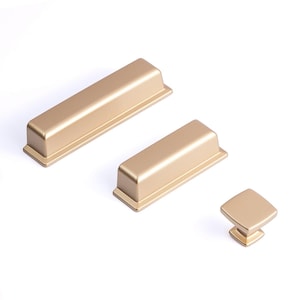 Op de afbeelding: Goudkleurige kastbeslagset, inclusief twee rechthoekige grepen en een vierkante knop. De hardware heeft een matte afwerking en een eenvoudig, modern ontwerp. De stukken zijn op een witte achtergrond geplaatst.