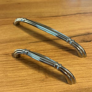 Pode incluir: Dois puxadores de armário em cromo polido com um design curvo e arqueado. Cada puxador apresenta um acabamento metálico liso e reflexivo com detalhes decorativos canelados perto dos pontos de montagem em ambas as extremidades.