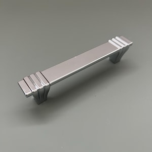 Può includere: Una maniglia per mobili argentata con una barra rettangolare e estremità decorative a gradini. L'hardware è realizzato in metallo lucido. La maniglia è lunga circa 12,7 cm.