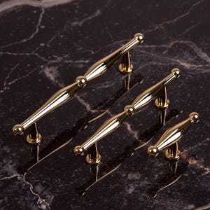 Peut inclure: Quatre poignées de meubles dorées au design classique. Chaque poignée présente un élément décoratif central et des extrémités arrondies. Les poignées sont présentées sur une surface sombre et marbrée, mettant en valeur leur finition polie.