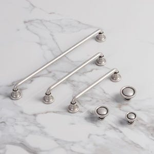Può includere: Una collezione di ferramenta per mobili color argento, tra cui tre maniglie a barra di diverse lunghezze e tre pomelli rotondi. La ferramenta ha una finitura anticata, che le conferisce un aspetto vintage. Gli articoli sono disposti su una superficie di marmo bianco.