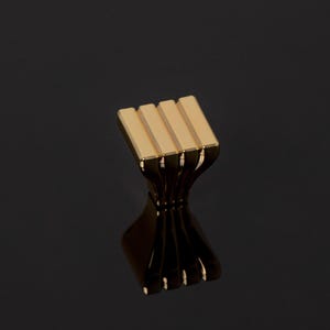 Può includere: Una maniglia dorata per armadi con una parte superiore rettangolare con quattro barre parallele. La base della maniglia si curva verso l'interno, quindi si allarga, creando un design decorativo unico. L'hardware è su uno sfondo scuro.