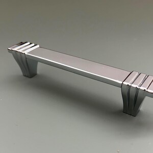 Puede incluir: Un tirador de armario cromado pulido con un centro rectangular y extremos decorativos. Los extremos presentan un diseño escalonado, añadiendo un toque Art Déco. Este herraje está diseñado para una fácil instalación y es adecuado para varios muebles.