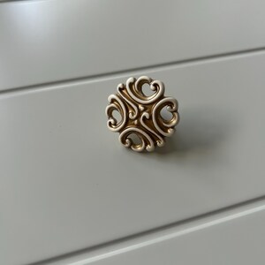 Può includere: Un pomello decorativo per armadi con un design a volute ornato. Il pomello presenta una finitura color oro con dettagli intricati, su uno sfondo grigio chiaro. Il design è simmetrico, con un punto centrale e curve radiali.