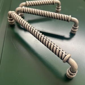 Puede incluir: Tres tiradores de armario de estilo antiguo con un diseño texturizado en forma de cuerda. Los herrajes metálicos tienen un acabado plateado y asas curvas. Los tiradores están dispuestos sobre una superficie verde oscuro.
