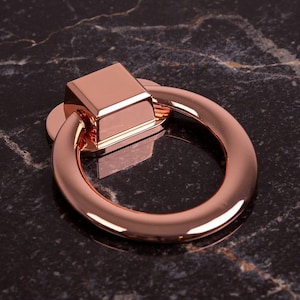 Może przedstawiać: Uchwyt do szafki w kolorze różowego złota z kwadratową górą i okrągłym uchwytem. Okucia umieszczone są na ciemnej, marmurowej powierzchni, odbijającej światło. Ten dekoracyjny uchwyt jest przeznaczony do mebli.