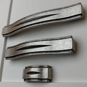 Könnte beinhalten: Drei antike Schrankbeschläge. Zwei sind gebogene, längliche Griffe, und einer ist ein kleinerer, rechteckiger Knopf. Alle sind in einem verwitterten Silberfarbton gehalten, mit einer strukturierten Oberfläche und einem zentralen Rillendesign.