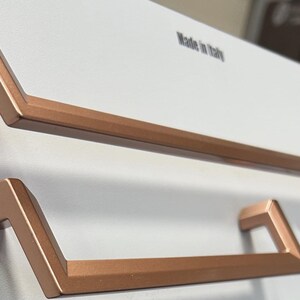 Op de afbeelding: Twee koperkleurige kastgrepen met een geometrisch ontwerp worden getoond tegen een witte achtergrond. De grepen zijn rechthoekig met schuine randen. De tekst "Made in Italy" is zichtbaar op de achtergrond.