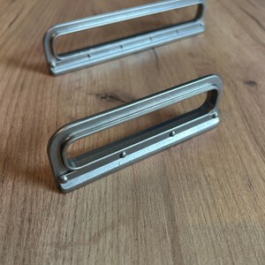 Può includere: Due maniglie per cassetti rettangolari in metallo color argento con un'apertura centrale liscia e allungata. Ogni maniglia presenta una base piatta con piccoli rivetti circolari lungo il bordo inferiore, offrendo un design pulito per mobili.