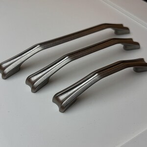 Peut inclure: Trois poignées de meubles en nickel brossé avec un design décoratif. Les poignées sont disposées en rangée, la plus grande en haut et la plus petite en bas. Les poignées ont une forme incurvée et sont en métal.