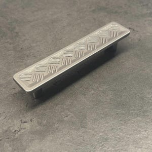 May include: A rectangular silver-toned metal drawer pull featuring a raised diamond plate tread pattern on its top surface. The handle has a smooth, flat border surrounding the textured center and includes two mounting posts on the underside.