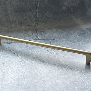 May include: A gold-colored cabinet pull with a straight bar and curved ends. The hardware is made of metal and is designed for use on drawers or cabinet doors. The pull is shown against a gray background.