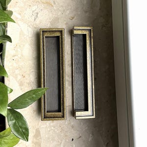 May include: Two rectangular cabinet pulls with a bronze finish. Each pull has a grooved, dark metal center and a decorative, raised border. The pulls are set against a light-colored, speckled surface.