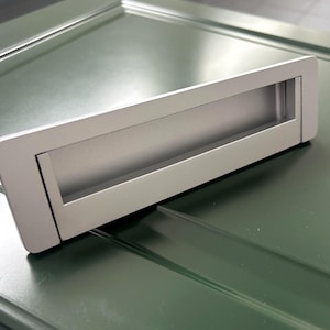 May include: A silver rectangular recessed door pull with a long, narrow opening. It's set against a dark green surface. The design is modern and minimalist, suitable for various interior styles. The pull is likely made of metal.