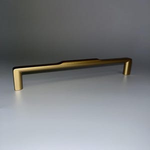 May include: A gold-colored cabinet pull with a rectangular bar and two vertical posts. The hardware is made of metal and has a matte finish. The pull is designed for easy gripping and opening of drawers or cabinets.