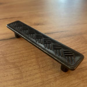 Puede incluir: Un tirador de cajón rectangular de metal con un acabado oscuro y desgastado, que presenta un patrón de relieve de chapa estriada en su superficie superior. El tirador se apoya en dos postes cilíndricos en la parte inferior para su fijación.