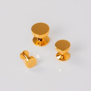 May include: Three gold-colored cabinet knobs with a round top and base, connected by a short, cylindrical stem. The knobs are arranged on a white surface, showcasing their reflective, polished finish. These decorative knobs are suitable for furniture.