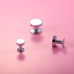 Könnte beinhalten: Drei silberne Schrankknöpfe mit weißen Oberseiten sind auf einer rosa Oberfläche angeordnet. Die Knöpfe haben einen zylindrischen Sockel und eine kreisförmige Oberseite. Ein Knopf liegt auf der Seite, während die anderen aufrecht stehen.