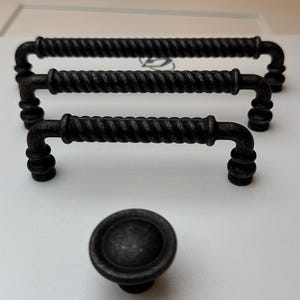 Op de afbeelding: Drie matzwarte metalen ladehandgrepen met een gedraaide touwtextuur en afgeronde uiteinden, gerangschikt in een getrapte formatie, vergezeld van een bijpassende ronde kastknop die op de voorgrond op een wit oppervlak is geplaatst.