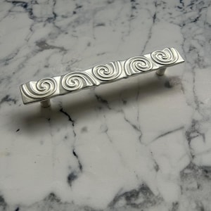 Puede incluir: Un tirador de armario plateado con una serie de diseños en espiral. El tirador rectangular tiene dos postes para el montaje. El herraje es de metal y está colocado sobre un fondo de mármol blanco y gris.