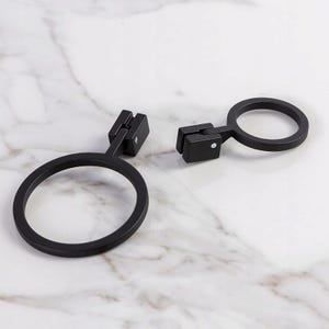 Op de afbeelding: Twee zwarte metalen ringvormige houders met een rechthoekige montagebeugel. De houders zijn ontworpen voor wandmontage. De achtergrond heeft een wit marmerpatroon.