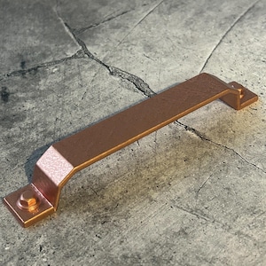 Può includere: Una maniglia per mobili color rame con una barra rettangolare e piastre di montaggio quadrate. L'hardware presenta una finitura strutturata ed è fissato con viti a vista. Questo pezzo è progettato per mobili e aggiornamenti di armadi.