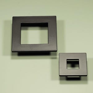 May include: Two square, dark bronze cabinet pulls with a recessed square center. The pulls are mounted on a light green background. One pull is larger than the other. Hardware for cabinets and drawers.