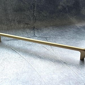 Pode incluir: Um puxador de armário dourado com uma barra reta e extremidades curvas. A ferragem é feita de metal e foi projetada para uso em gavetas ou portas de armários. O puxador é mostrado em um fundo cinza.