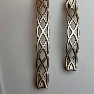 Puede incluir: Dos tiradores metálicos para armarios con un diseño entrelazado. El tirador más grande mide unos 15 cm de largo, y el más pequeño unos 10 cm. Diseño decorativo e intrincado.