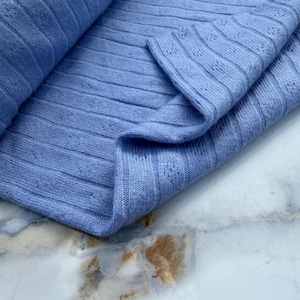 May include: A light blue knit fabric featuring a vertical ribbed texture with small, repeating eyelet perforations between the ribs. The material is folded over itself, showcasing the soft, consistent weave and the uniform periwinkle blue color.