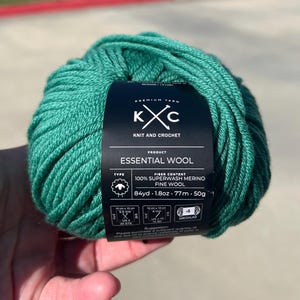 May include: A ball of teal Essential Wool yarn. The label reads "KXC Knit and Crochet" and "100% Superwash Merino Fine Wool." The yarn is 77m, 50g. The label also indicates a medium weight. The ball of yarn is being held.