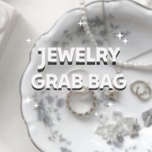 May include: A white, floral-patterned dish with the text "JEWELRY GRAB BAG" in bold, white letters. The dish contains various pieces of jewellery, including rings, earrings, and a pearl necklace. The jewellery is silver-toned.