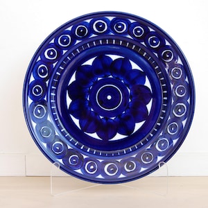 May include: A round ceramic plate featuring a deep blue and white color scheme. The center displays a floral-inspired pattern surrounded by a ring of circular motifs. The outer edge is defined by a solid blue border, creating a bold contrast.