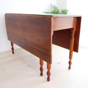 May include: A wooden drop leaf table with a dark brown finish and turned legs. The table has a rectangular top with two drop leaves that can be raised or lowered.