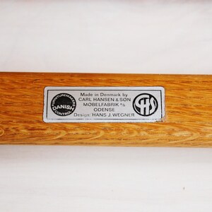 May include: Close-up of a wooden furniture component with a rectangular label. The label reads "Made in Denmark by CARL HANSEN & SON MOBELFABRIK ODENSE Design: HANS J. WEGNER" and includes a "Danish Control" seal.