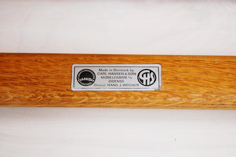 May include: Close-up of a wooden furniture component with a rectangular label. The label reads "Made in Denmark by CARL HANSEN & SON MOBELFABRIK ODENSE Design: HANS J. WEGNER" and includes a "Danish Control" seal.