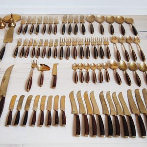 May include: A collection of vintage flatware, including forks, spoons, and knives, with brass-colored metal and dark brown wooden handles. The set includes serving pieces and salt and pepper shakers.