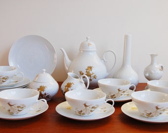 Puede incluir: Un juego de té de porcelana blanca con detalles dorados. El juego incluye una tetera, una jarra de crema, un azucarero y seis tazas de té y platillos. Las tazas de té y los platillos tienen un delicado diseño floral con detalles dorados.