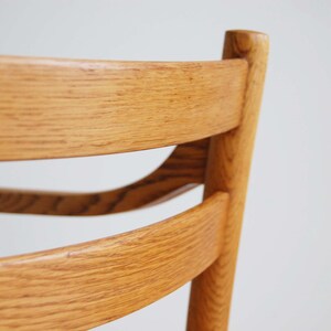 May include: Close-up of a wooden chair back. The chair is made of light-colored wood with a visible grain. The design features curved back supports and a vertical post, showcasing a classic furniture style.
